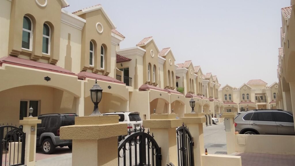 Row of Mediterranean-style townhouses and villas in a residential community, likely The Meadows, Dubai, featuring light beige/yellow walls, red roof accents, arched windows, and covered carports. The image shows a quiet street with parked cars and black metal gates.
